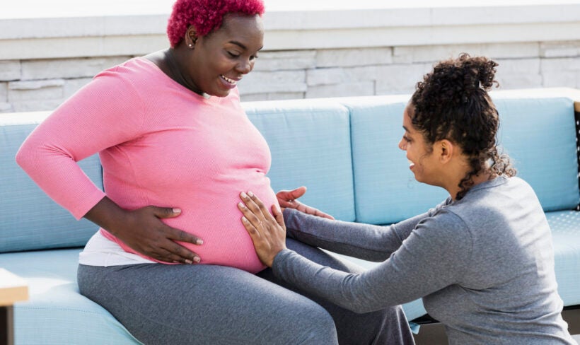 A pregnant African-American woman with her doula or birth support coach. The expectant mother, who had pink hair and is wearing a pink shirt, is sitting outdoors on a patio sofa smiling. The doula, a mixed race woman, is kneeling in front of her, touching her round abdomen. Both women are in their 30s.