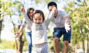 A happy toddler learning to walk in a park with her smiling parents behind