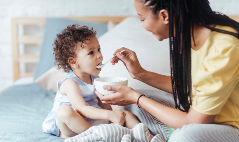 A smiling mother using a spoon to feed her toddler